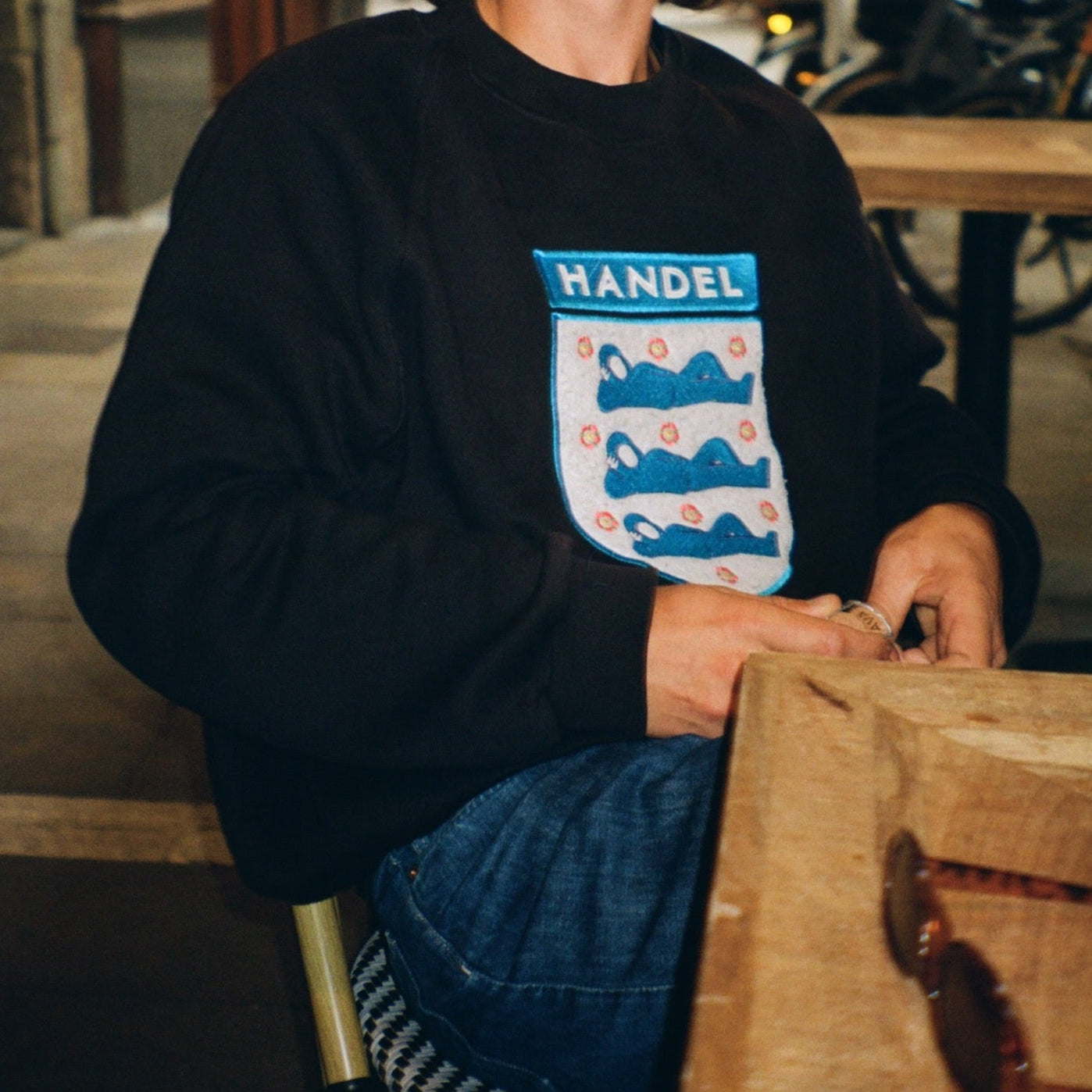 Person wearing a black Handel sweatshirt featuring a stylish embroidered crest logo with blue figures and floral details, captured in a relaxed café setting. Modern design meets high-quality craftsmanship.