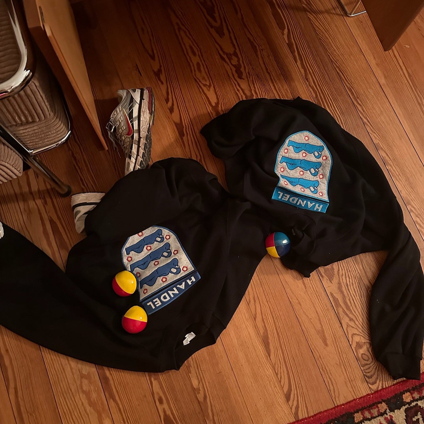 two black Handel sweatshirts featuring embroidered crest logos with blue figures and floral details, placed on a wooden floor alongside juggling balls and sneakers. A casual and artistic display of modern craftsmanship